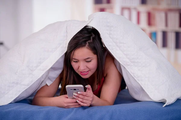 Gros plan d'une femme souriante avec sa tête sous son oreiller à l'aide de son téléphone portable, essayant de dormir la nuit, concept d'insomnie — Photo