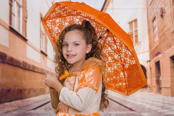 Güzel sömürge kostüm giyen ve bulanık bir arka planda turuncu bir şemsiye tutan küçük güzel kızın yakın çekim — Stok fotoğraf