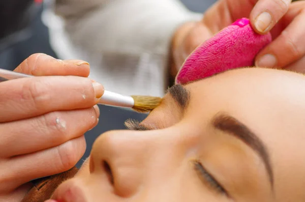 Porträtt av vackra ansikte ung kvinna att få make-up. Konstnären ansöker eyeshadow på hennes ögonbryn med borste. Damen blundade med avkoppling, i en suddig bakgrund — Stockfoto