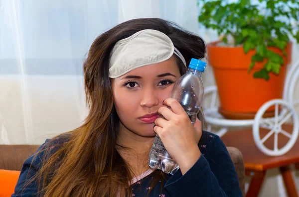 Μεθυσμένος γυναίκα με μια μάσκα ματιών ύπνου στο κεφάλι της και με ένα μπουκάλι νερό πατώντας στο πρόσωπό της, χρησιμοποιώντας την άλλη πλευρά, έννοια πονοκέφαλο — Φωτογραφία Αρχείου