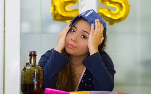 Mujer joven aplicando una bolsa de hielo en su cabeza usando ambas manos y una botella de alcohol en la mesa, concepto de resaca — Foto de Stock