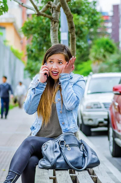 Moda modny casual młoda kobieta ubrana jean kurtkę i czarne legginsy, za pomocą jej telefon komórkowy i pokazując szok emocje i dotykając jej twarz z jej strony. Siedzi na zewnątrz, portret kobiety z — Zdjęcie stockowe