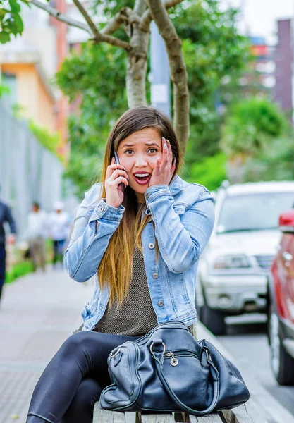 Cep telefonunu kullanarak ve şok duygu gösterilen ve eliyle yüzünü dokunaklı bir jean ceket ve siyah tayt giyen moda trendy rahat genç kadın. Açık havada, kadın portre ile oturan — Stok fotoğraf