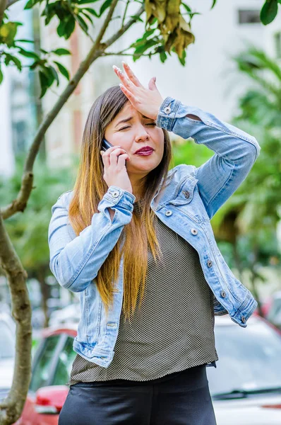 Fashion trendy casual young woman wearing a jean jacket and black leggings, using her cellphone and showing shock emotion with her hand in the head. Female portrait with shocked facial expression — Stock Photo, Image