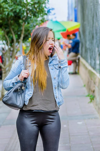 Bulanık bir arka planda bir jean ceket ve siyah tayt giyen sokaklarda yürüyüş ve uzun bir gün sırasında esneme moda trendy rahat genç kadın — Stok fotoğraf