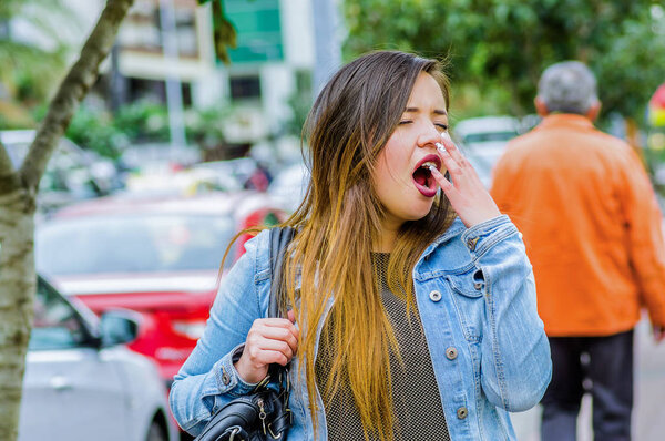 Fashion trendy casual young woman walking in the streets and yawning during a long day, wearing a jean jacket and black leggings in a blurred background