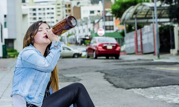 Bella giovane donna ubriaca seduta su un marciapiede con una bottiglia di birra in mano e bere, donna esperta e concetto di tossicodipendente — Foto Stock