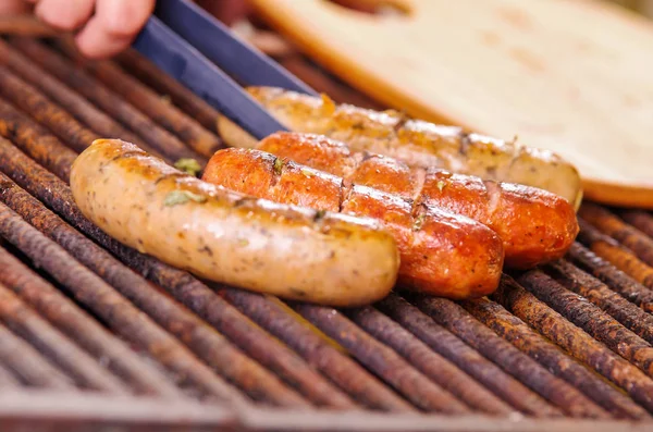 Primer plano de la mano de la mujer sosteniendo unas pinzas girando las salchichas a la parrilla en la parrilla de barbacoa. Barbacoa. Embutidos bávaros — Foto de Stock