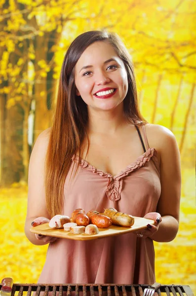 Primer plano de sonriente hermosa mujer joven sosteniendo en sus manos una salchichas a la parrilla en la tabla de cortar de madera, barbacoa en el jardín. Embutidos bávaros —  Fotos de Stock