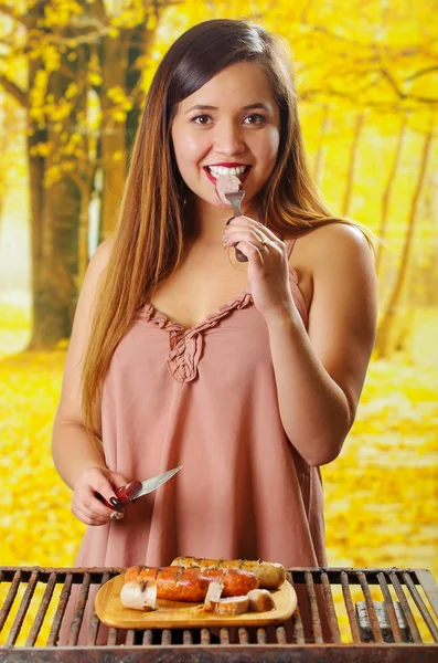 Close up of smiling beautiful young woman eating a piece of sausage using a fork and knife in her hands, and grilled sausages on wooden cutting board, BBQ in the garden. Bavarian sausages, in a — Stock Photo, Image