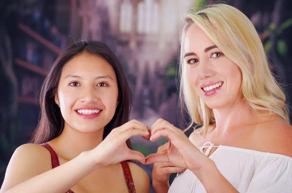 Two young women blonde and latin girl smiling and breaking racism idiosyncrasy from a american person and foreign people, both doing a heart sign with teir hands, racism, violence or discrimination