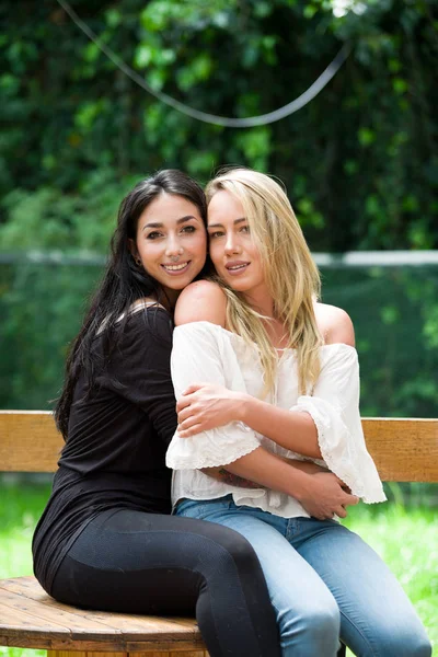 Um par de lésbicas orgulhosas ao ar livre sentadas em uma mesa de madeira, mulher morena está abraçando uma mulher loira, em um fundo de jardim. — Fotografia de Stock