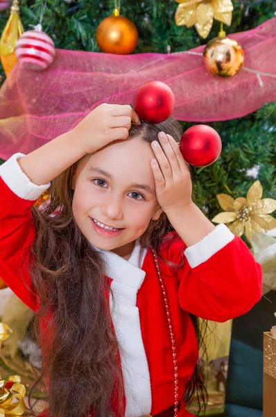 Close up van lachende meisje draagt een rode santa kostuum en twee kerstballen bedrijf in haar handen en poseren over haar hoofd, met een kerstboom achter, Kerstmis concept — Stockfoto