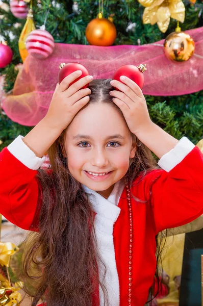 Primer plano de niña sonriente con un disfraz de santa roja y sosteniendo dos bolas de Navidad en sus manos y posando sobre su cabeza, con un árbol de Navidad detrás, concepto de Navidad — Foto de Stock