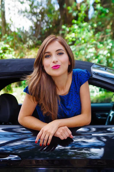 Close up de mulher bonita vestindo um vestido azul e posando dentro de um carro preto de luxo em uma viagem. o carro de pé nas laterais, em um fundo de natureza turva — Fotografia de Stock