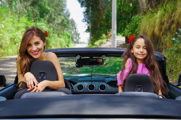 Close up of beautiful woman wearing a black dress and a red flower in her head and her gorgeous daugher a pink dress with a red flower in her head and posing inside of a luxury black car on a road — Stock Photo, Image