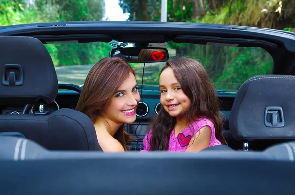 Close up of beautiful woman and her gorgeous daugher wearing a pink dress posing inside of a luxury black car and looking at camera on a road trip, in a blurred nature background — Stock Photo, Image