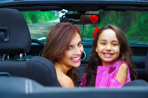 Close up of beautiful woman and her gorgeous daugher wearing a pink dress posing inside of a luxury black car and looking at camera on a road trip, in a blurred nature background — Stock Photo, Image
