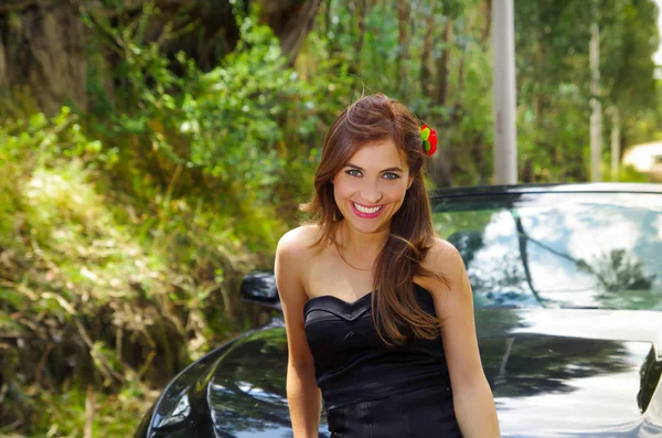 Close-up van de mooie vrouw het dragen van een zwarte jurk en een rode bloem in haar hoofd en poseren voor een zwarte luxeauto op een road trip, de auto staande op de zijlijn, in een wazig natuur — Stockfoto