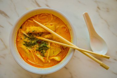 Penang asam laksa pirinç şehriye kase içinde yemek çubukları ile beyaz kase görünümünü baharatlı balık demirhindi çorba ve beyaz porselen kaşık ve Singapur taze otlar ile beyaz tabloda yukarıda