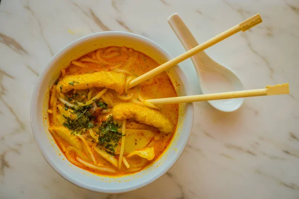 Vista superior de penang asam laksa fideos de arroz en tazón blanco con palillos dentro del tazón en la mesa blanca con pescado picante sopa de tamarindo y cuchara de porcelana blanca y hierbas frescas en Singapur — Foto de Stock