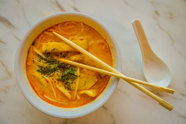 Vue ci-dessus de penang asam laksa nouilles de riz dans un bol blanc avec des baguettes à l'intérieur du bol sur la table blanche avec soupe de tamarin de poisson épicé et cuillère en porcelaine blanche et des herbes fraîches à Singapour — Photo