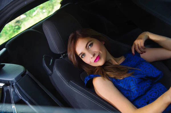 Close up of beautiful woman wearing a blue dress in a reclined seat and posing inside of a luxury black car on a roadtrip, the car standing on the sidelines, in a blurred nature background — Stock Photo, Image