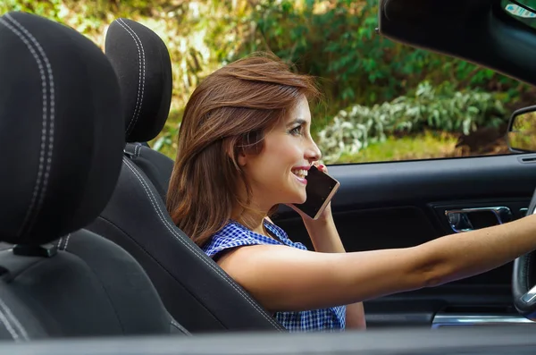 Крупним планом красива молода жінка використовує свій мобільний телефон всередині чорного автомобіля, керуючи її машиною однією рукою, на розмитому фоні природи — стокове фото
