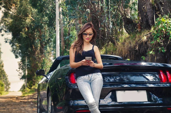 Gros plan de belle femme caucasienne utilisant son téléphone portable et portant des lunettes de soleil, t-shirt noir et jeans, la voiture debout sur les lignes de touche — Photo
