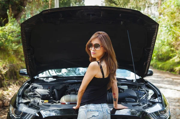 Piękne kobiety sobie sunglases, czarny t-shirt i dżinsy, problem z czarnym samochodem na roadtrip, pozowanie na kamery i dając kopię, samochód, stojący na uboczu., kaptur z bliska — Zdjęcie stockowe