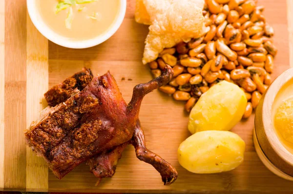 Cut pieces cooked guine pig meat lying on wooden surface next to potatoes, tostados and bowl of salsa — Stock Photo, Image