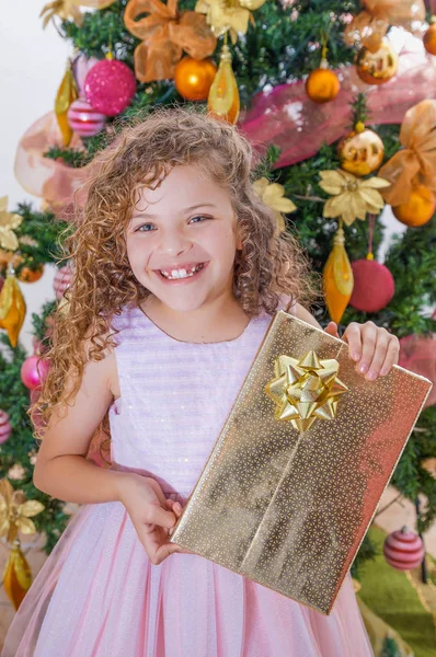 Primer plano de niña rizada sonriente con un vestido rosa y sosteniendo un gran regalo en sus manos, con un árbol de Navidad detrás, Navidad y el concepto de año nuevo — Foto de Stock