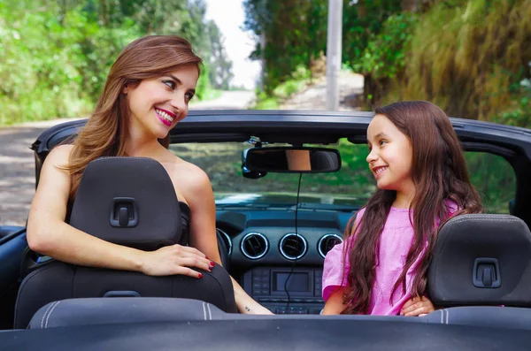 Close up of beautiful woman wearing a black dress and a red flower in her head and her gorgeous daugher a pink dress with a red flower in her head and posing inside of a luxury black car on a road — Stock Photo, Image