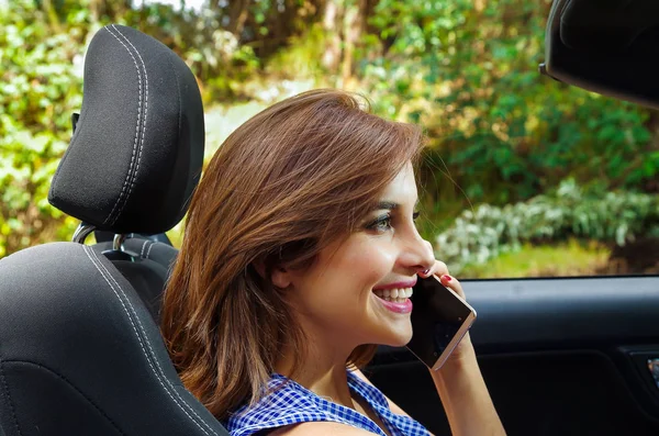 Porträtt av vackra kaukasiska kvinna via hennes mobil insidan svart bilen, samtidigt som hon driver i en suddig natur bakgrund — Stockfoto