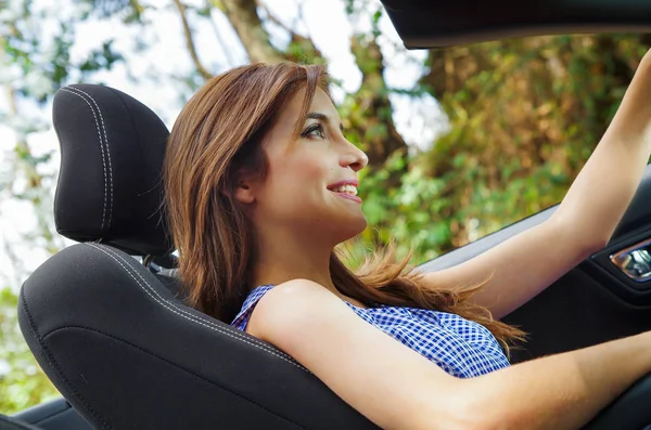 Under beskåda av vacker ung kvinna inne i en svart bil, kör sin bil med en hand, i en suddig natur bakgrund — Stockfoto