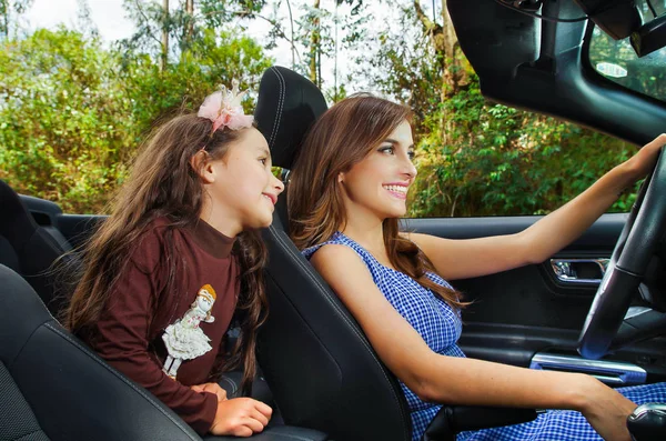 Below view of beautiful young woman with her daugher inside of the black car, driving her car with one hand, in a blurred nature background — Stock Photo, Image