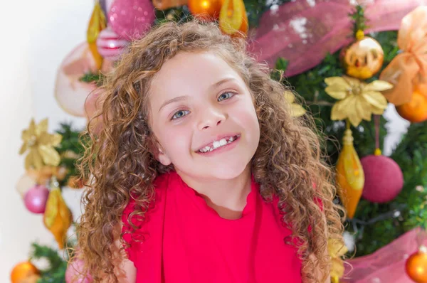 笑顔の美しい巻き毛の少女は赤いブラウスを着て、クリスマス ツリー、クリスマスと新年の概念の背後にあるカメラのポーズのクローズ アップ — ストック写真