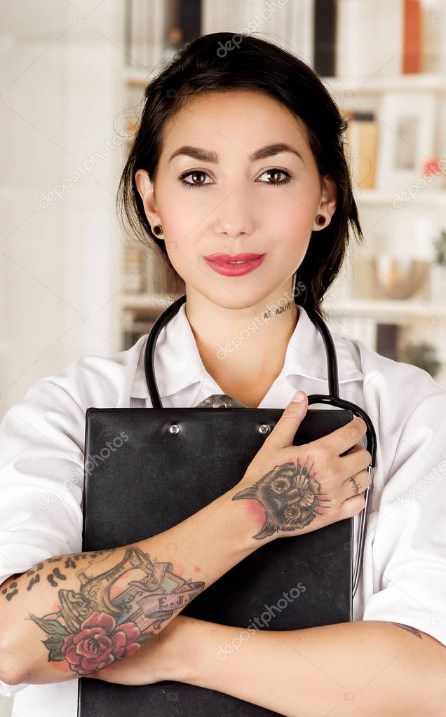 Beautiful tattooed young doctor hugging a black binder in office background