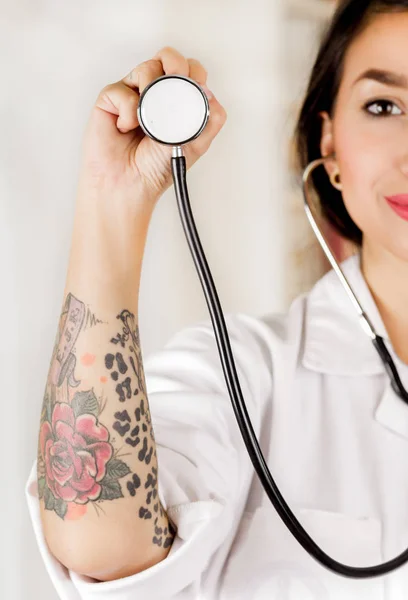 Beautiful tattooed young doctor holding the chest part of the stethoscope in her hand, in office background — Stock Photo, Image