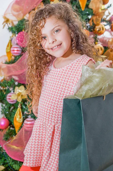 Gros plan de fille bouclée souriante tenant un énorme sac de cadeau, avec un arbre de Noël derrière, Noël et concept de nouvelle année — Photo