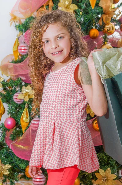 Gros plan de fille bouclée souriante tenant un énorme sac de cadeau, avec un arbre de Noël derrière, Noël et concept de nouvelle année — Photo