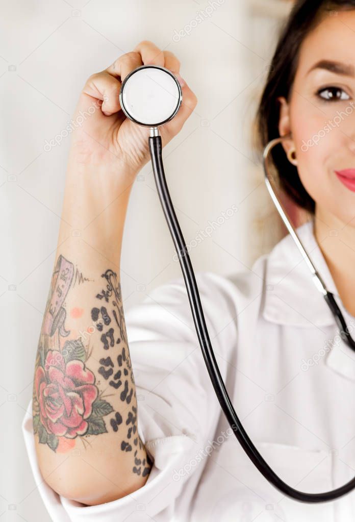 Beautiful tattooed young doctor holding the chest part of the stethoscope in her hand, in office background