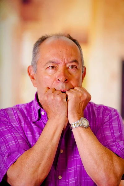 Ritratto di un uomo nervoso maturo, con entrambe le mani in bocca, che indossa una t-shirt quadrata viola su sfondo sfocato — Foto Stock