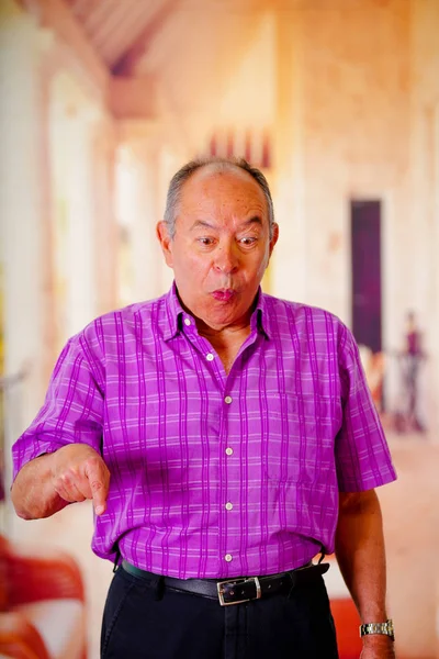 Крупным планом счастливого старика, показывающего рукой вниз и одетого в фиолетовую квадратную футболку на размытом фоне — стоковое фото