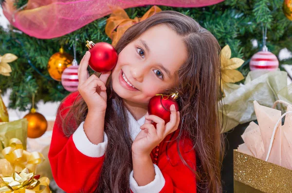 Close up of happy girl wearing a red santa costume and holding two christmas balls in her hands and pressing over her face, with a Christmas tree behind, christmas concept — Stock Photo, Image