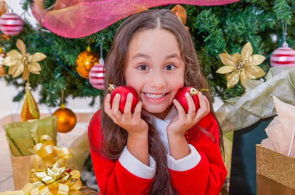 Close up of happy girl wearing a red santa costume and holding two christmas balls in her hands and pressing over her face, with a Christmas tree behind, christmas concept — Stock Photo, Image
