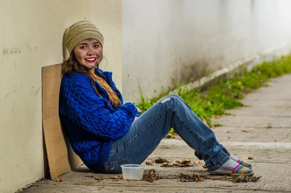 Außenansicht einer obdachlosen lächelnden Frau, die bei kaltem Herbstwetter auf der Straße bettelt und auf dem Fußboden auf dem Bürgersteig sitzt — Stockfoto