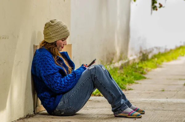 Außenansicht einer obdachlosen Frau, die bei kaltem Herbstwetter auf der Straße bettelt und mit dem Handy in der Hand auf dem Fußboden auf dem Bürgersteig sitzt — Stockfoto