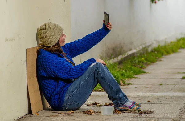 Vista al aire libre de la mujer sin hogar mendigando en la calle en el frío clima otoñal sentada en el suelo en la acera tomando una selfie —  Fotos de Stock