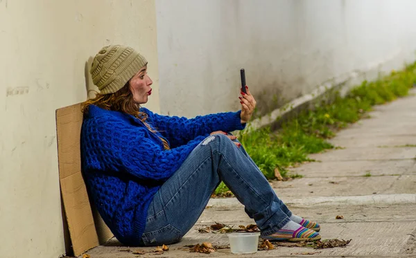 Vista ao ar livre da mulher sem-teto implorando na rua em tempo frio outono sentado no chão na calçada com um celular em sua mão — Fotografia de Stock